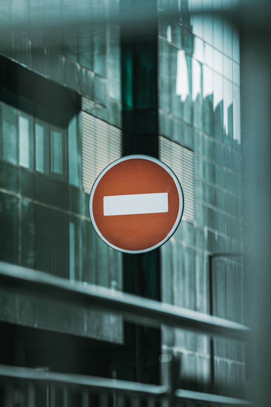 A red no entry sign in front of a modern glass building on a city street.